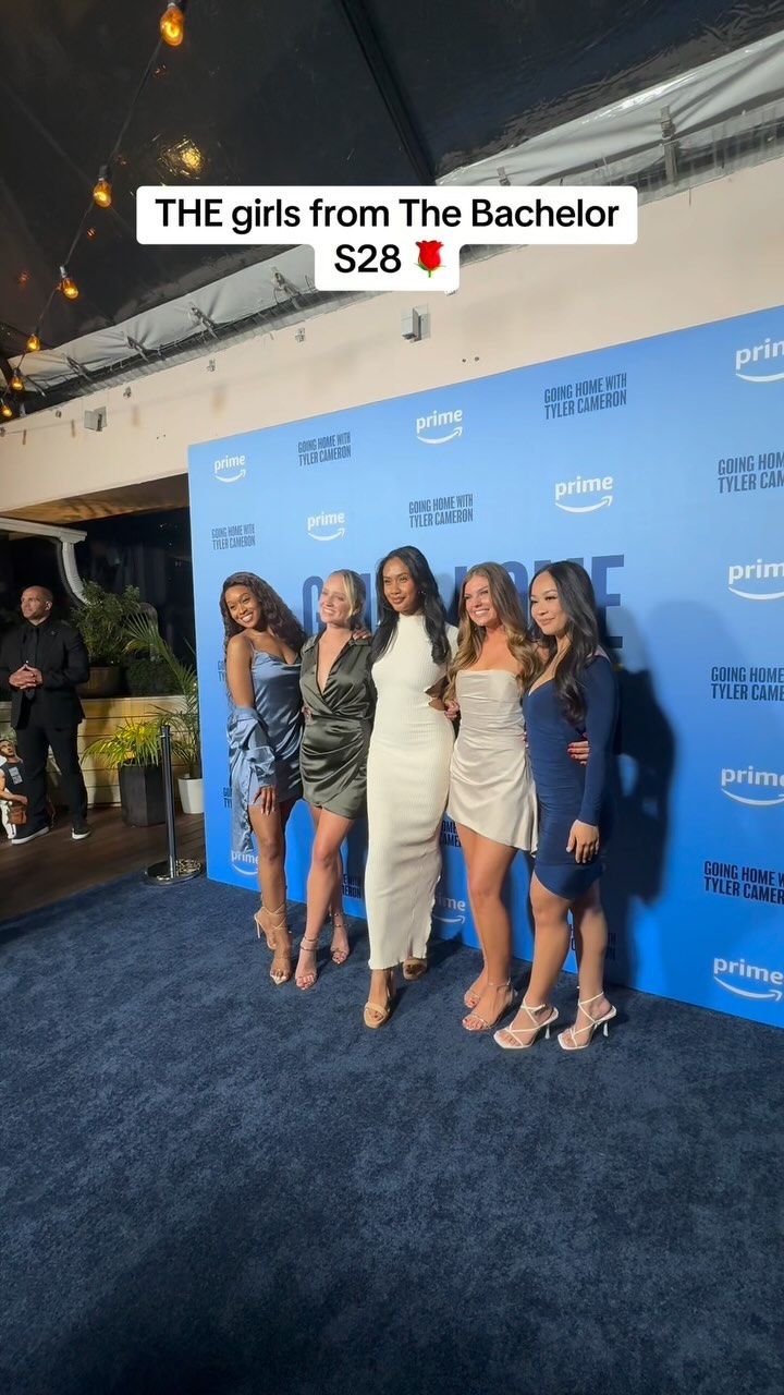 A group of six women posing on a blue carpet in front of a backdrop with the Prime Video logo and text. They are dressed in various outfits, from short dresses to a long white gown, and are smiling and looking at the camera. The lighting is bright, and there are strings of lights overhead.