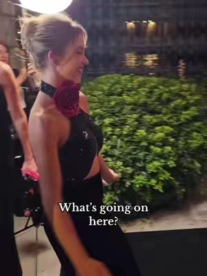 A woman in a black dress walks towards the camera with a posed expression. She has her hair up, a choker with a large rose detail, and appears to be on a red carpet or at an event. The background is slightly blurred, suggesting a busy setting.