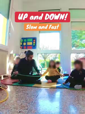 A group of children and a man are sitting on a colorful floor mat, each holding a hula hoop. The man is in the center, guiding the children in a musical activity. Above, text overlays read 'UP and DOWN!' and 'Slow and Fast'. The background is a bright room with large windows.