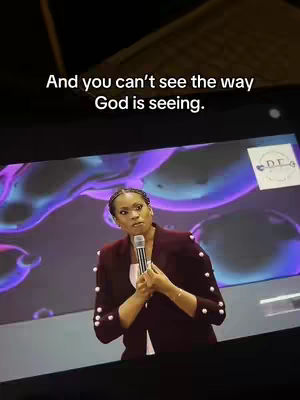 The image shows a woman speaking and holding a microphone on a stage with a large video screen in the background. She's wearing a maroon blazer and a white shirt, and the background has abstract, purple and blue shapes, with some text indicating its from D.E. The text overlay at the top says, "And you can't see the way God is seeing."