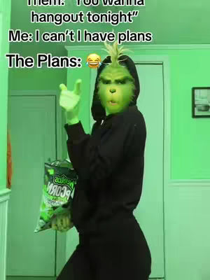 A person in a Grinch mask and costume is holding a bag of popcorn snacks in a green-lit room. The person is wearing a black hoodie and has green skin, a Grinch face mask, and green gloves. Text overlays read, "Them: You wanna hangout tonight", "Me: I can't I have plans", and "The Plans: 🤣".
