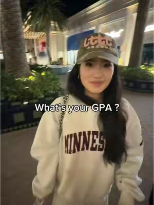 A young woman, wearing a hat and smiling, stands in an outdoor setting at night. She has long dark hair and is wearing a white hoodie with the word "MINNES" in large maroon letters across the front. The background shows lit-up storefronts and palm trees, suggesting an urban or commercial area.