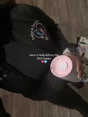The video features a person from the shoulders down, dressed in black clothing. They hold a clear, round object that glows with a pale pink light., over a wooden-panelled surface. A logo displaying "Labor & Delive" on the left breast of their top. The text "omw to help deliver the 2025 babies" appears as an overlay with two hearts.