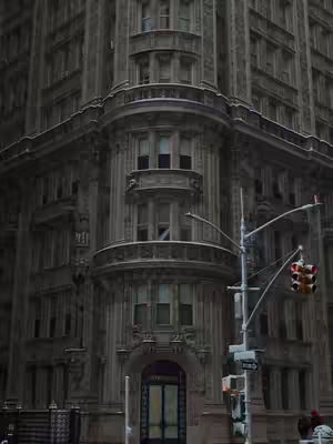 A photo of a large, ornate building in New York City. The building is made of dark gray stone and has many windows and balconies. The sky is overcast. A building corner is on the left and the right. Traffic lights hangs on the right.