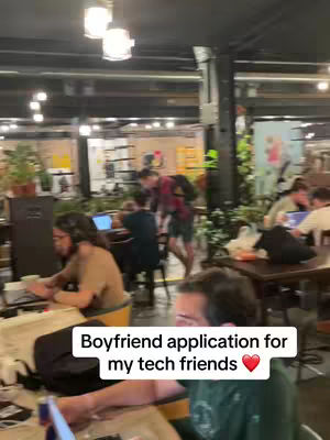The cover image shows a bustling indoor scene, likely a cafe or open-plan office, filled with people working on laptops and socializing. The lighting is warm, and the setting appears relaxed and collaborative. Text overlayed at the bottom reads 'Boyfriend application for my tech friends ❤️'.