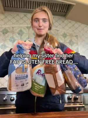 A young woman with blonde hair smiling and holding various bags of gluten-free bread products. She is in a kitchen with a stainless steel hood, and tiled walls.