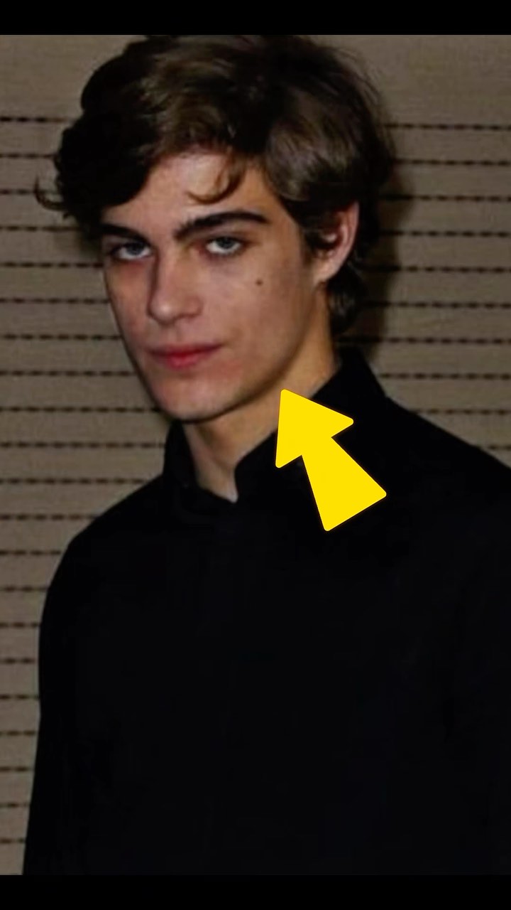 A close-up shot of a young man with dark hair and a serious expression. He is wearing a black shirt, and the background has a striped pattern. A yellow arrow points to his neck. The lighting appears to be soft.