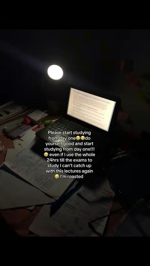 A dimly lit scene featuring a laptop displaying study notes. The laptop sits on a cluttered desk with scattered papers and a round light source overhead. The image has a dark, warm tone.