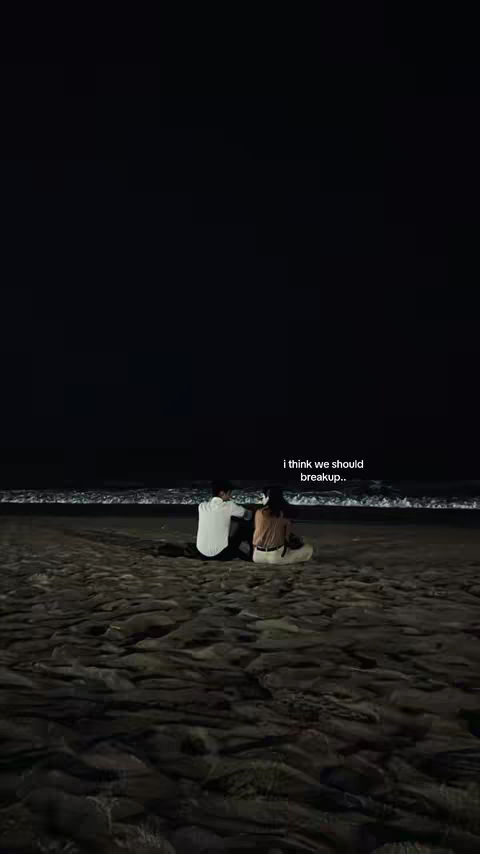 The image shows a couple sitting on a sandy beach at night, with the ocean in the background. The scene is dark overall, with the night sky above and the water visible along the horizon. The couple is silhouetted as they sit closely together, with soft lighting reflecting off the water.