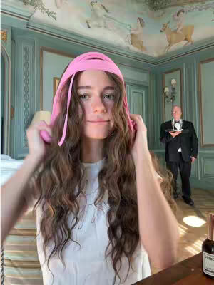 A young woman with long wavy brown hair is the central focus, wearing a white top and a pink head wrap. She's holding the head wrap, positioned as if adjusting it. The background suggests a high-ceilinged room with ornate wall paneling in light green, adorned with painted murals and a chandelier. A man in formal attire, possibly a butler, stands near the far right, adding a sense of formality to the scene. Additionally, a bottle of wine is visible on the desk on right side of the woman.