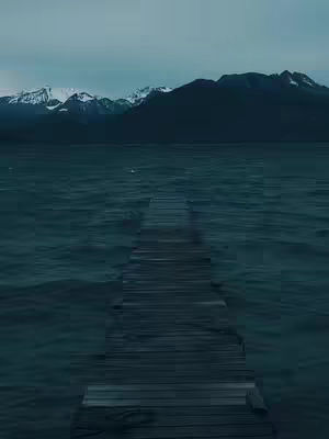 An eye-level shot of a wooden pier extending out into a dark teal ocean. In the background, dark silhouettes of mountains can be seen, some with white snow caps under a cloudy, gray sky. The image has a cool, muted color palette.