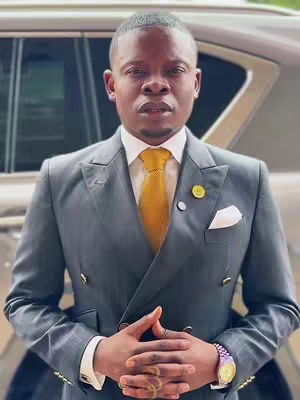 A man in a formal suit posing in front of a car. He has a serious expression, hands clasped, and wears a tie and accessories.