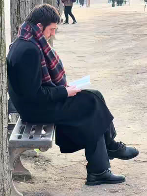 A man in a winter coat and scarf sits on a park bench, reading a book. The background is blurred, suggesting a park setting with other people visible.