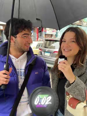 A couple standing outside under an umbrella. The woman smiles and holds a microphone. The man holds the umbrella and looks at the woman. Behind them are cars and buildings.