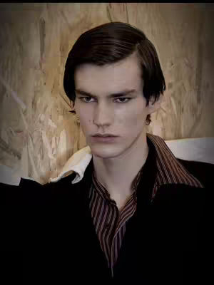 Close-up shot of a young man with dark hair and a serious expression. He's wearing a striped shirt under a black jacket, with a light-colored wooden panel background.