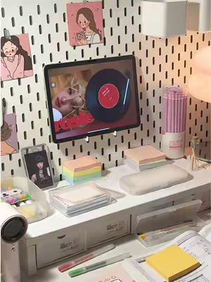 A well-lit, aesthetic workspace featuring a white desk, a pegboard wall with decorative elements, and various stationery items. A tablet displaying a music app interface dominates the scene, with a record-like visual. The color palette is light and pastel-toned.
