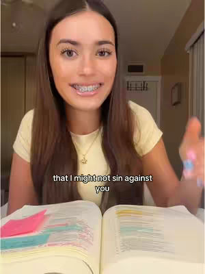A young woman with long brown hair and braces is centered in the frame, smiling and looking directly at the camera. She wears a yellow shirt and a necklace. In front of her is an open book with colorful bookmarks. A text overlay appears at the bottom of the image, and the overall composition suggests a personal and engaging video aimed at viewers.