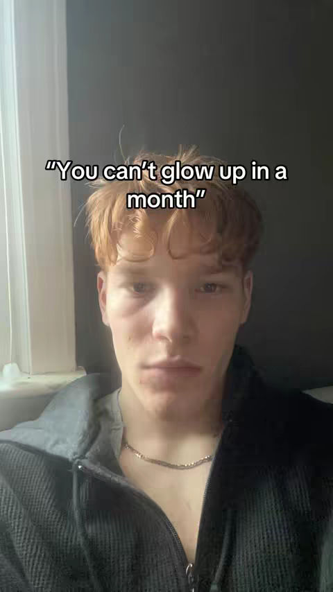 A close-up shot of a young man with light skin and reddish-brown hair, looking directly at the camera. The background is a blurred, neutral gray. The man is wearing a black hooded jacket and a silver necklace.
