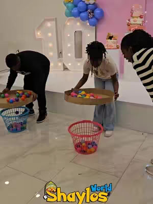 The cover image shows a party game environment, likely a birthday party, with three children interacting. The background features a large decorative number 10, balloons, and a cake. The children are actively involved in a game, each holding a tray or basket with colorful balls, with other baskets on the floor. The setting is brightly lit with a polished floor. The logo of 'World Shaylos' appears at the bottom.
