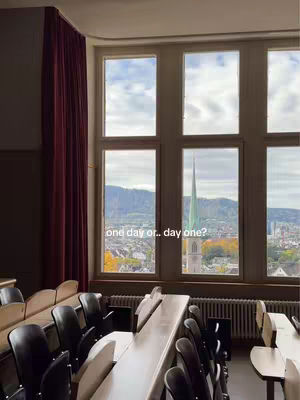 The image captures a view from a classroom, showcasing a large window offering a scenic view of a city and mountains. Inside, there are rows of desks and chairs, suggesting a lecture hall setting. The image is well-lit, featuring a color palette dominated by natural light and the muted tones of the interior, creating a calm atmosphere.