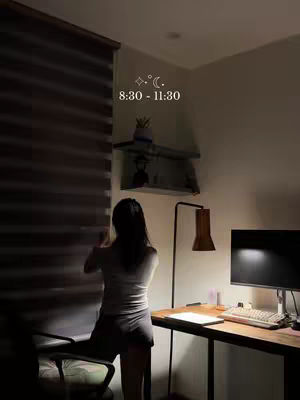 A dimly lit room with a person standing by a window and desk area. The person is viewed from behind, partially obscured by the scene's shadows. A desk with a computer and lamp are in the lower right. The image has a warm, muted color palette.