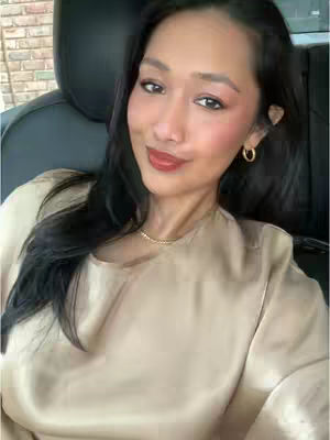 A woman with long black hair is seated inside a car. She is wearing a beige top, a gold necklace, and small gold hoop earrings. Her makeup includes a red lipstick. The car interior is dark, and a brick wall is visible in the background. She has a warm and inviting expression.