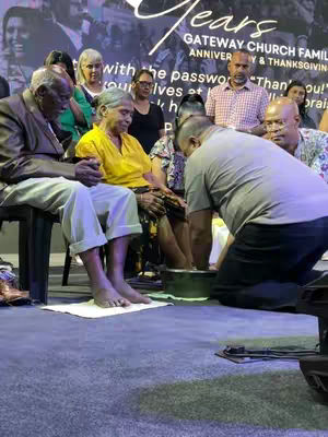 The image shows a group of people in a church setting, appearing to be a celebration. In the foreground, an elderly man and woman sit on chairs, their feet in a basin of water, while another person kneels to wash their feet. Other individuals are in the background. The color palette is dominated by the blue-gray carpet, the pale tones of those depicted, and the dark basin of water, with a backdrop featuring commemorative text or a church banner.