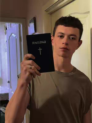 A young man holds up a black book with a cross symbol and the letter 'R N N N N' on the cover. He stands indoors, likely in a home, with a neutral expression, suggesting a message he is conveying.