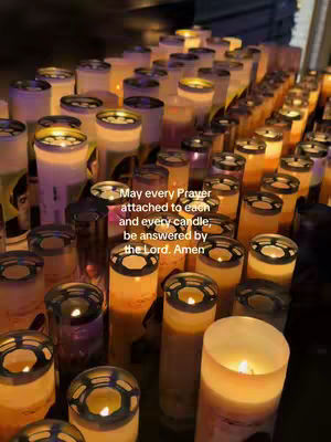An image featuring a low-angle shot of numerous lit candles, arranged closely together. The candles are cylindrical, with a blend of translucent and opaque white or beige wax, and feature a metal or metallic-looking cap. The flames are visible, casting a warm glow. The background is dimly lit, emphasizing the candles' light. Text overlays stating a prayer is placed over the candles.