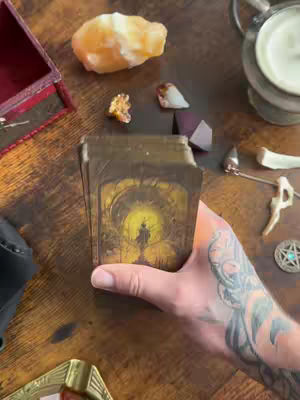 An overhead shot displays a wooden surface scattered with various mystical items. A hand, heavily tattooed with a detailed design, firmly holds a deck of tarot cards, the back of a card featuring a silhouette within a circular design. Surrounding the hand and cards are an orange crystal, small decorative objects, a silver chalice, and small crystals adding to the mystical aesthetic.