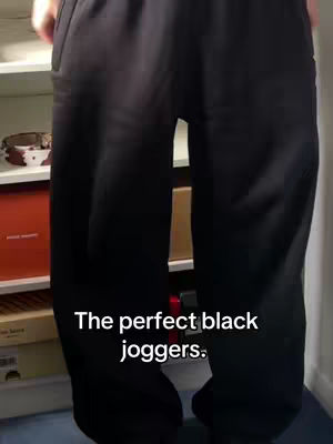 The image shows a person wearing black joggers. The person is standing, their torso and legs filling most of the frame. The joggers are the focal point, and the background is a plain wall and shelves with visible items.