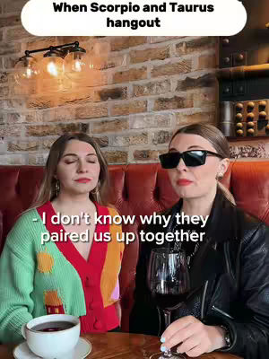 Two women are seated, facing the camera in a dimly lit area. An overhead banner is visible with text, and one woman is holding a glass of wine. The background features a brick wall and lighting fixtures.