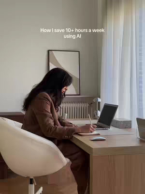 A woman in a brown suit is seated at a desk, appearing to work on a laptop and write. The setting is a well-lit room with a window, curtains, and a neutral color palette.