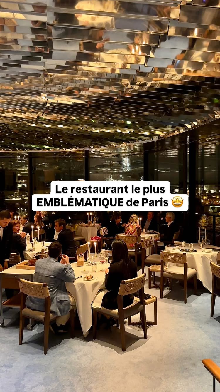 The image shows the interior of a high-end restaurant, with numerous patrons seated at tables. The ceiling is covered with mirrored panels reflecting the lights and surroundings. Tables are set with white tablecloths, silverware, and candles. People are dressed in formal attire. The overall composition is elegant and luxurious.