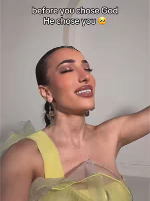 A woman with dark hair and makeup is smiling with her eyes closed, framed from the chest up. Her hair is pulled back and she is wearing a yellow dress. There is text overlayed at the top of the image.