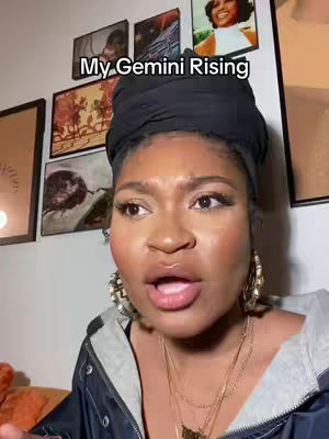 A woman with dark skin and a black head covering looks directly at the camera with an open mouth, as if speaking. Behind her are framed pictures on a wall, and the text overlay reads 'My Gemini Rising.' The overall composition focuses on the woman's face, framed by a dark head wrap, with a neutral background.