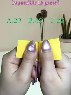 The image shows a close-up view of hands holding a small, yellow notebook shaped like a book. The nail polish is a reflective, pearlescent pink color. Behind the hands, are some flowers in a clear vase, options A.23, B.32, C.22 in green font labeled above the hands and the words "Impossible to guess?" in pink font. The background has a cream or beige color.