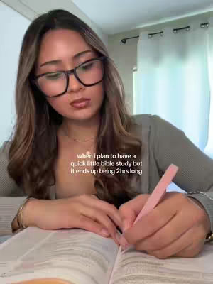 A young woman with long brown hair and glasses is sitting at a desk, presumably studying. She is holding a pink pen and writing in a large book that appears to be a bible. The background is blurred and includes hints of a window with a curtain.