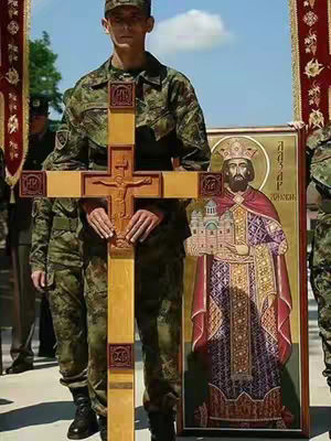 A man in a military uniform stands holding a large wooden cross, with an icon depicting a religious figure on his right. Other people in military uniforms stand in the background, next to a banner with red and gold decorations. The sky is clear and blue.