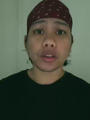A close-up shot of a person facing the camera directly, wearing a burgundy patterned bandana that covers the head, a black t-shirt. The background is a plain, off-white wall. The lighting is moderate, and the person appears to be speaking.