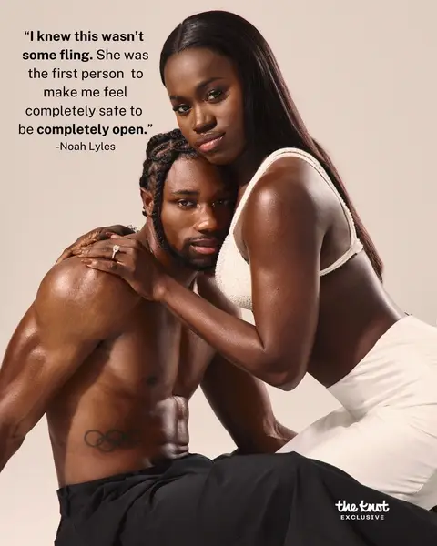 A romantic image showing a man and a woman embracing. The man, with short, styled hair, is shirtless with an olympic rings tattoo on his upper back and is looking at the camera.  The woman is wearing a white top and white pants, and has her arm around the man with her head leaning towards his. The background is a pale neutral color.