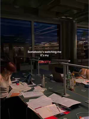 A dimly lit scene depicts a study area in a library or similar space. In the foreground, a person, presumed to be the content creator, is visible, illuminated by the warm light of a desk lamp. Papers and a laptop litter the workspace, and the person appears to be studying. The background reveals a large glass window offering a view of an urban cityscape lit by streetlights against the dusk sky. Text overlayed on the image reads, "Somebody's watching me it's my ANXIETY".