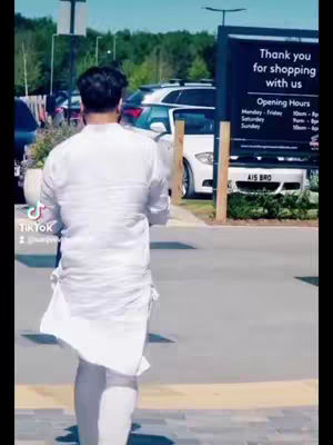 A man in a white outfit is seen from behind, walking away from the camera. The background includes a parking lot with cars, a sign that says 'Thank you for shopping with us,' and some trees under a blue sky. The overall composition is a daylight shot.