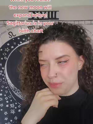 A young woman with curly brown hair gazes to the side, her expression thoughtful. She's wearing a black top and has subtle makeup. Behind her is a tapestry with celestial designs.
