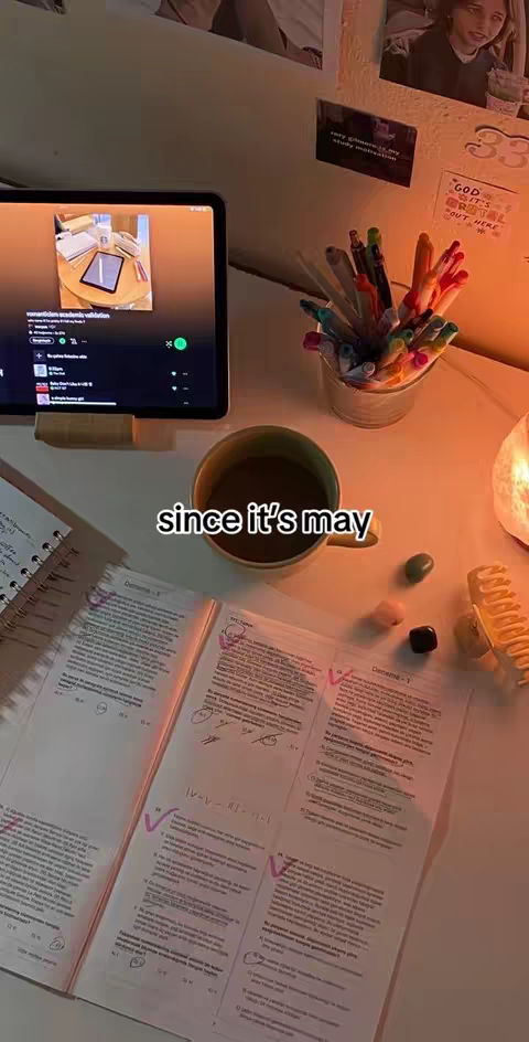 A desk with schoolwork, a coffee mug, and an open laptop displaying a music app. Text overlayed reads 'since it's may'. The scene is warmly lit.