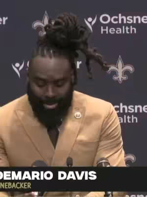 An African American man in a tan suit jacket is speaking into a microphone. He has his hair up in a bun and is wearing a blazer. The background includes a banner with the logos of a health company and the Saints team.