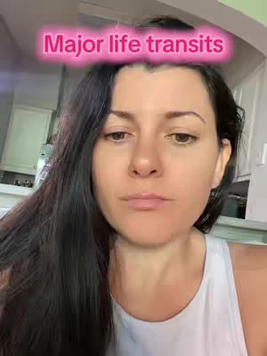 A woman with dark hair is centered in a selfie-style shot. Above her head, a text overlay reads 'Major life transits' in a pink, outlined font. The background is blurred, but hints at a kitchen setting.