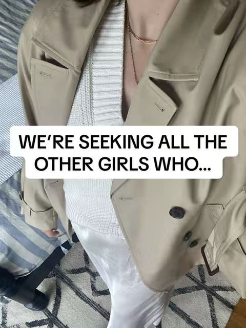 The cover image features a person, likely female, from the chest up, wearing a beige trench coat over a white sweater and dress. The person has a golden necklace and a portion of a black boot is visible. The background is blurred, featuring a bedspread and a patterned rug. The coat is the focal point, with the white sweater and dress providing contrast. Text overlayed on the image reads: 'WE'RE SEEKING ALL THE OTHER GIRLS WHO...'.