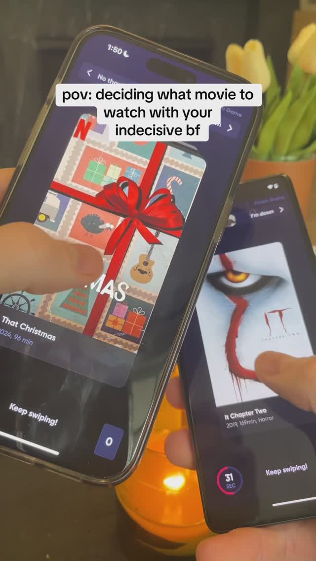 Two smartphones are held in hands, displaying movie app interfaces. One shows a festive Christmas-themed poster, the other a poster for 'It Chapter Two.' The background includes a blurred setting with flowers and a glowing candle.