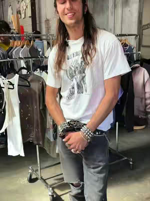 A man with long hair and a white t-shirt stands in front of racks of clothing. He is smiling and looking at the camera. He is wearing silver bracelets and rings, accessorized with a belt. In the background, several racks of clothing are visible.